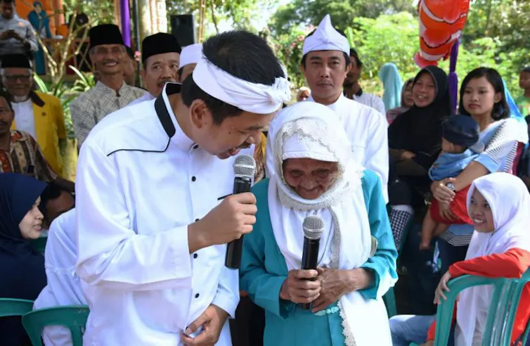 Dedi Mulyadi mengenakan iket Sunda berbicara dengan warga sambil dikelilingi kerumunan yang memperhatikannya.