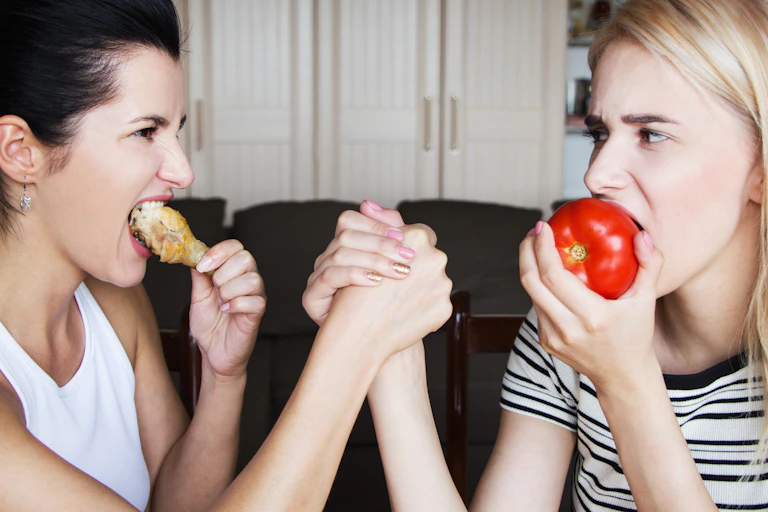 Two women arm-wrestling. One eats a chicken leg and one easts a beef tomato