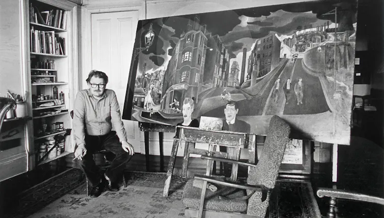 A black and white image of the artist and writer Alasdair Gray with one of his paintings in the living room of his Glasgow flat.