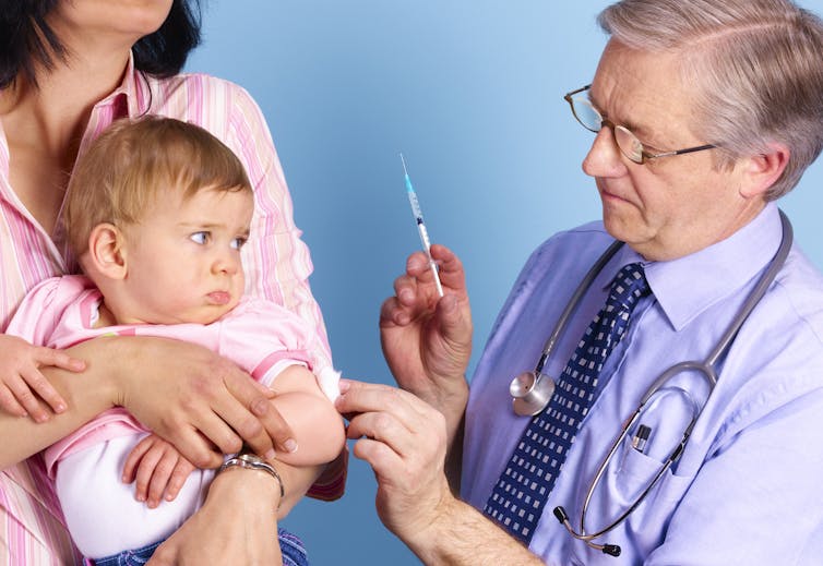 Grumpy white baby giving side-eye to an older white male doctor about to administer a vaccine