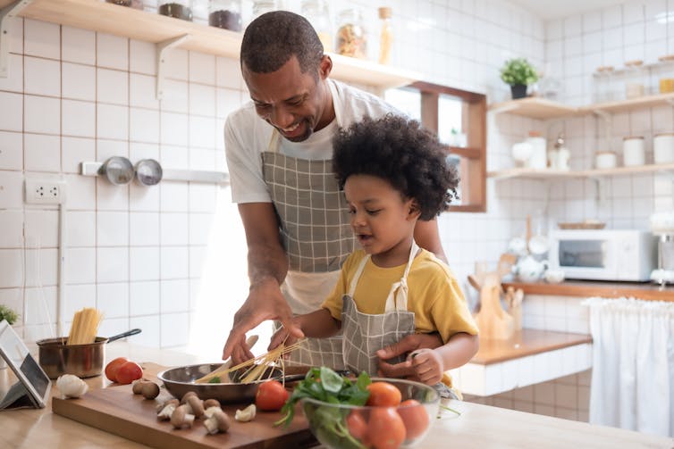 A father stands behind a child at a stove helping them with a stir fry.