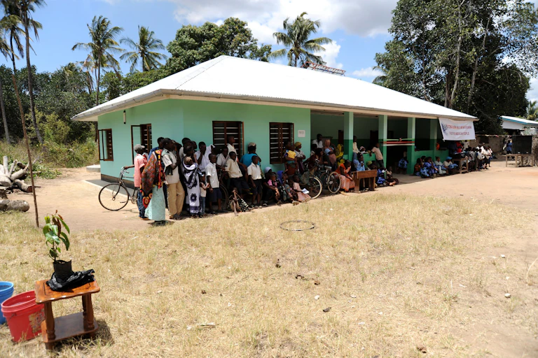 People outside a single storey building, some with bicycles. Palm trees in the background.