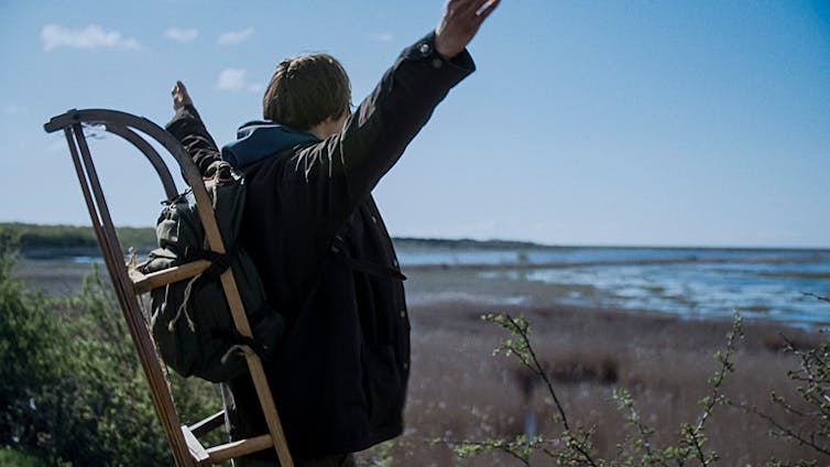 young man arms up looking at flooding coast with rucksack and sledge on back