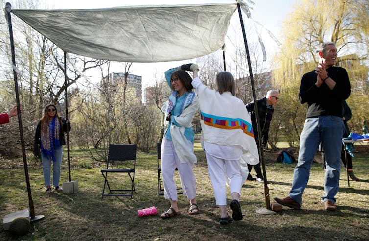 Two women wearing white clothes and prayer shawls dance under a simple canopy in a park as a few people look on.