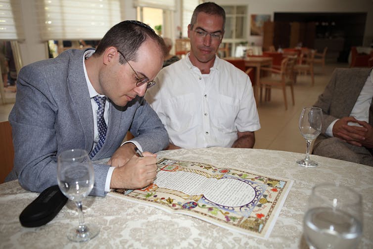 A man in a blue-gray suit signs a colorfully decorated piece of paper as another man in a white shirt watches.