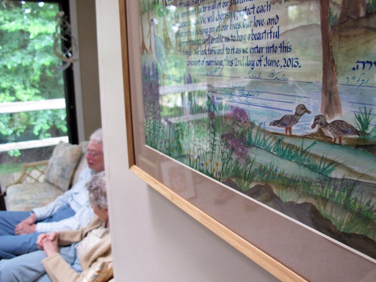 A colorful, framed drawing on a white wall, with two older women barely visible sitting on a couch at the back of the room.