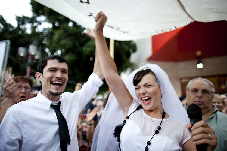 A man and woman, both wearing white, smile as they raise their joined hands above their heads.