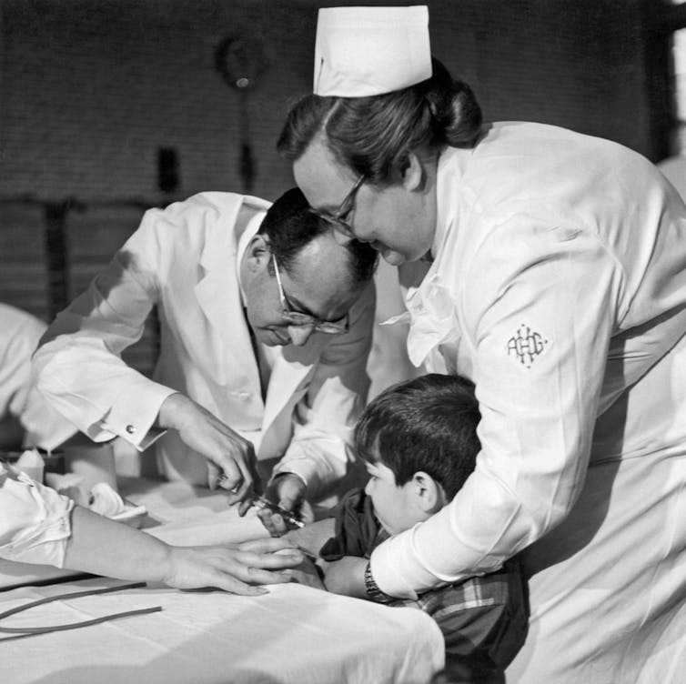 A black-and-white photo of a doctor in a white coat giving an injection to a boy who is held by a female nurse.