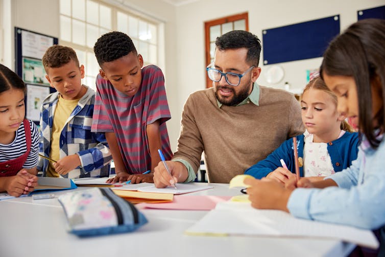 a man sitting at a desk in a classroom, young kids stand around him as he explains something