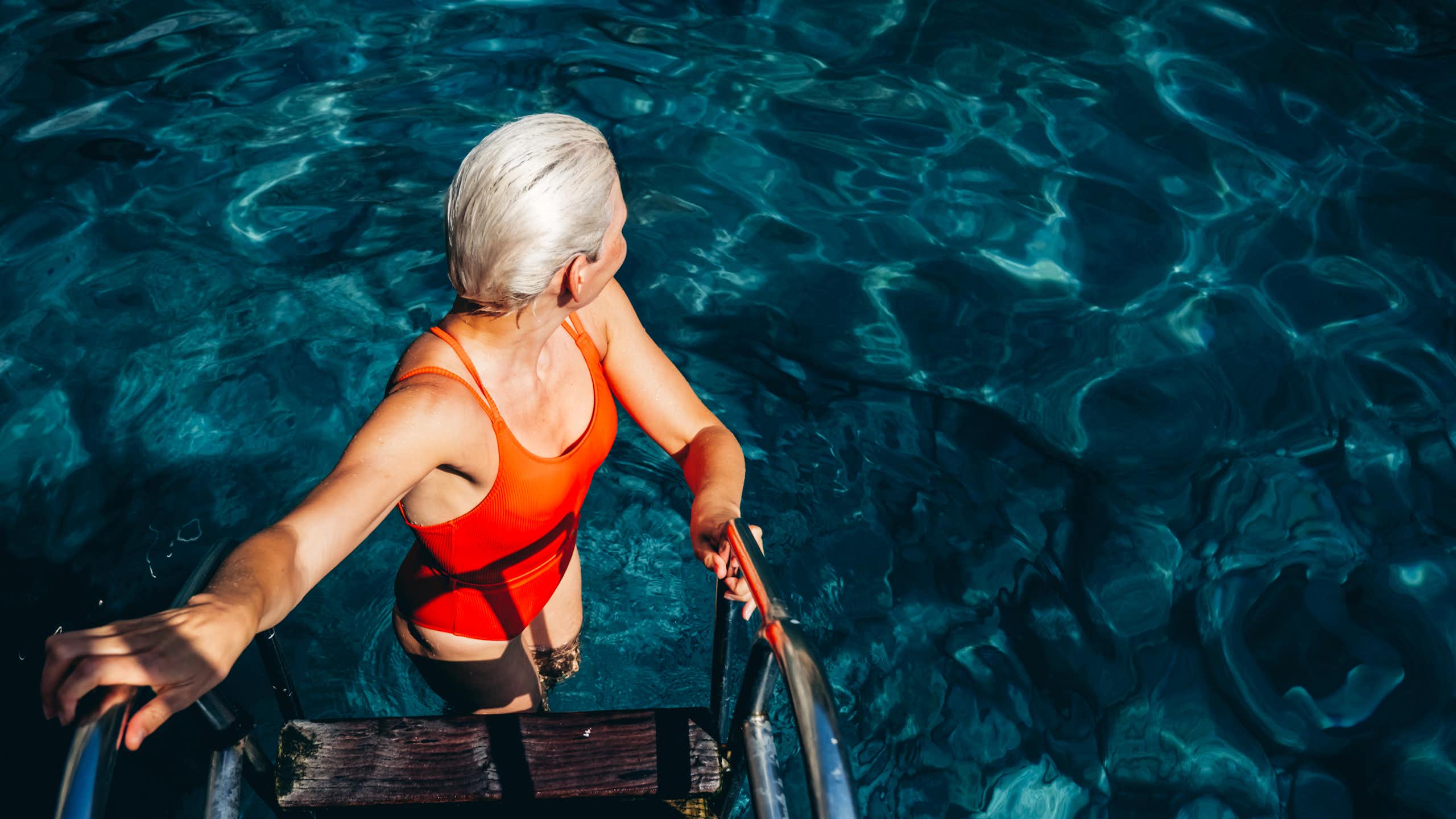 Une femme aux cheveux blancs et en maillot de bain se tient dans une échelle de piscine