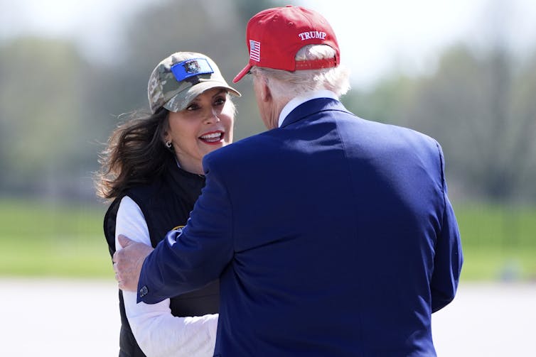 A woman shakes hands with a man in a blue suit wearing a red ballcap.
