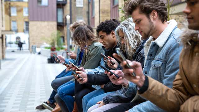 eight young adults sit in a row in a city plaza looking at their phones