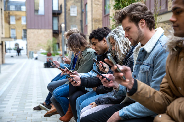 eight young adults sit in a row in a city plaza looking at their phones