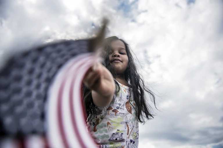 a Black girl holds a small flag near the camera amid a cloudy sky