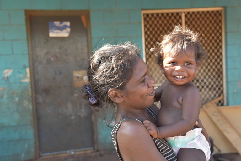 A young Aboriginal woman holds a toddler in her arms in front of a house