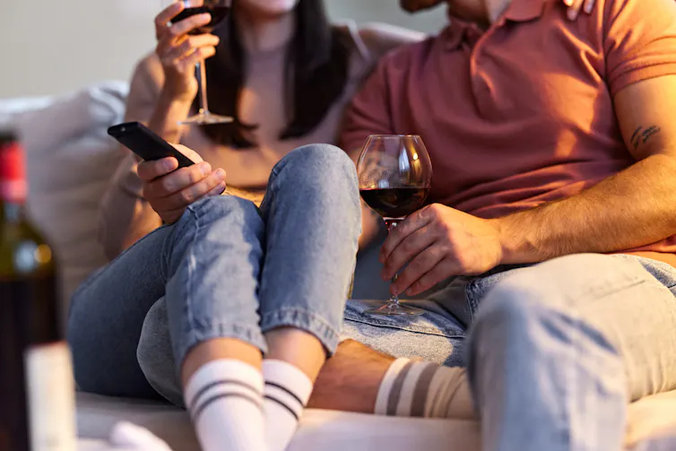 Bodies of man and woman sitting on a couch with tv remote and glasses of wine.