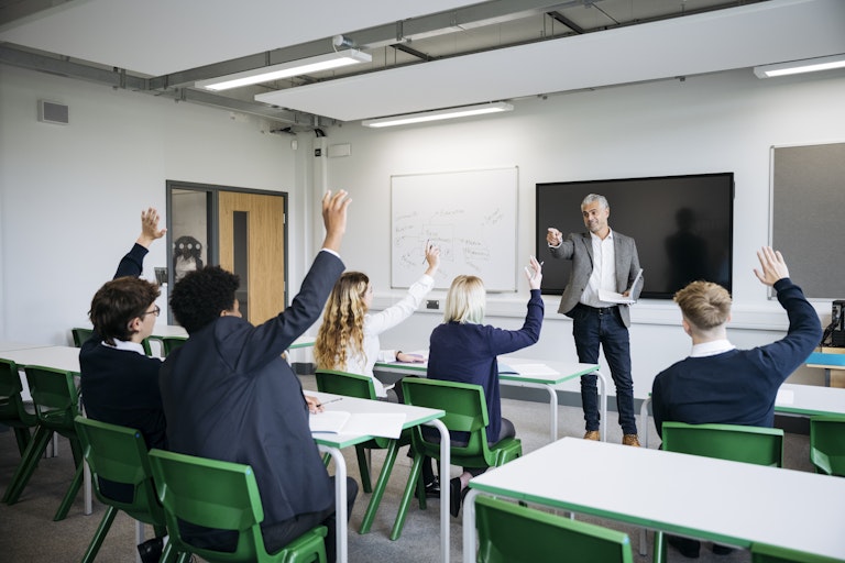 A teacher stands at the front of the classroom, pointing to students who have their hands raised.