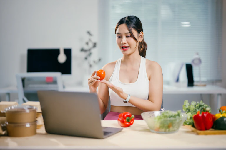 Une jeune est face à son écran d'ordinateur en train de composer une salade, elle a une tomate à la main.