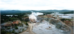 A giant area where the earth has been removed and mining is taking place with smoke going up from a bomb