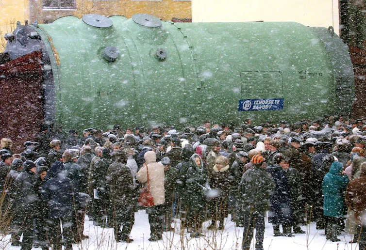 A crowd of people stand in the snow in front of a large cylindrical reactor body.