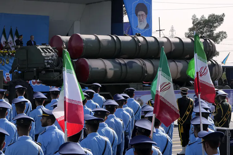 Men in powder-blue uniforms and carrying green, white and red Iranian flags march in front of a large piece of military equipment.