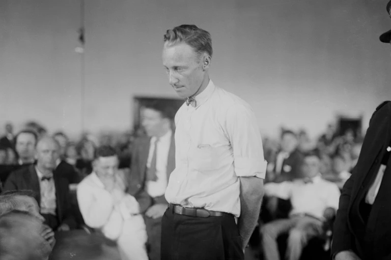 A man in a white shirt looks downcast as he stands among rows of seated people.