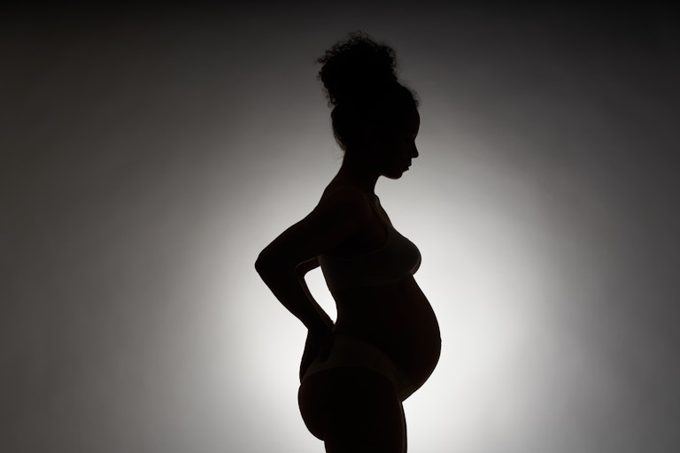 A photo of a silhouette in profile of a young woman, with curly hair in a bump, stands and holds her hands on her back.