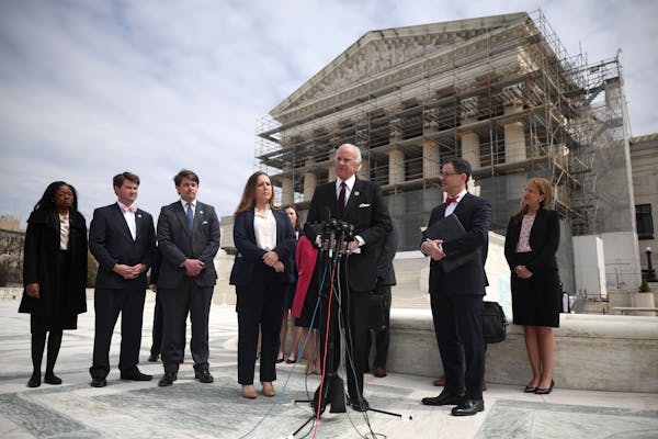 Man in suit speaks into a microphone, flanked by other people who are standing in front of a building surrounded by scaffolding.