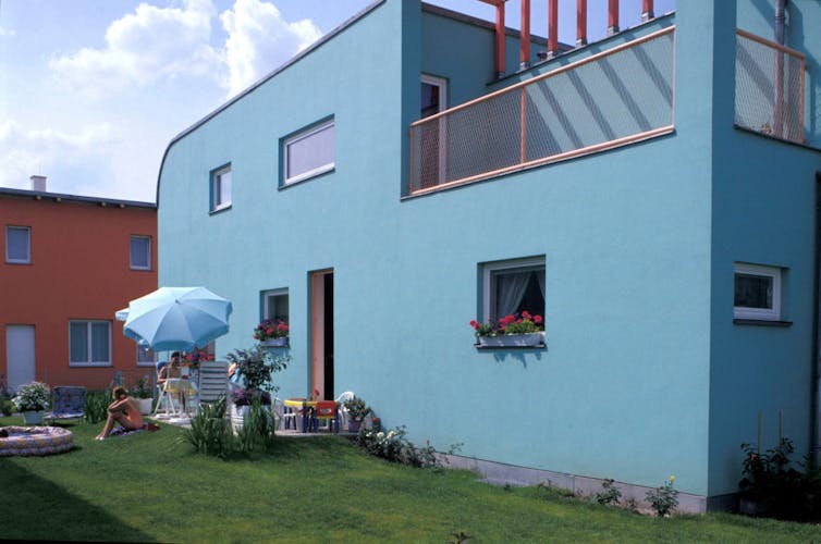 A blue, modern-looking, two-story dwelling with red flowers in the windows.