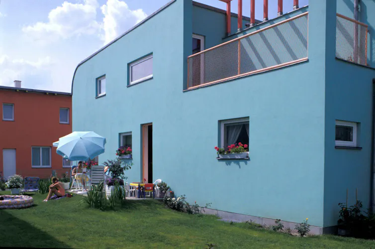 A blue, modern-looking, two-story dwelling with red flowers in the windows.