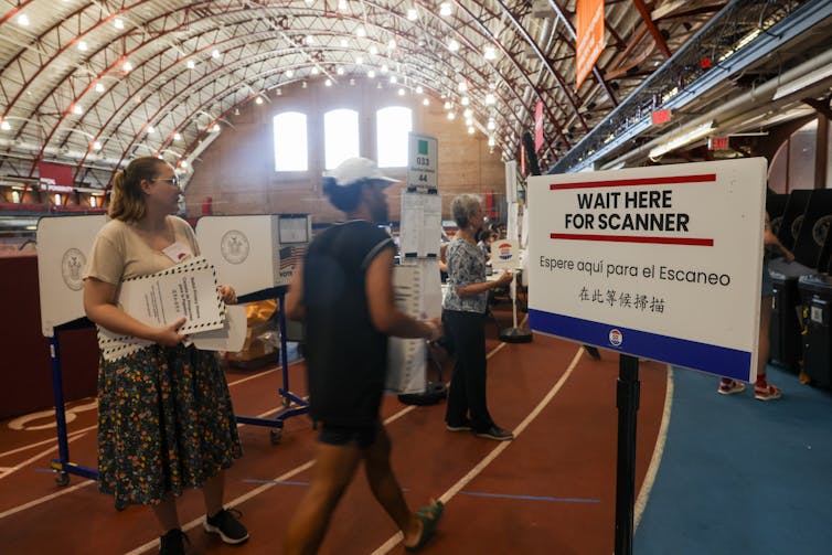 Several people are seen moving around white voting booths and holding white folders in a large room.