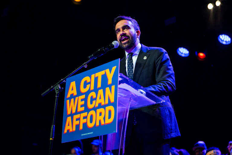 A man with a beard and a dark suit and tie stands as a podium that has a placard on it that says, 'A city we can afford.'