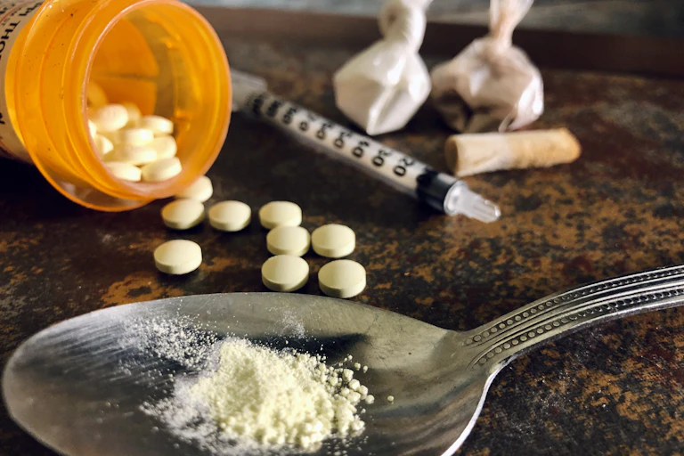 Two small sacks of cocaine, a syringe, cocaine on a spoon and opioids spilling out of a pill bottle sit on a table.
