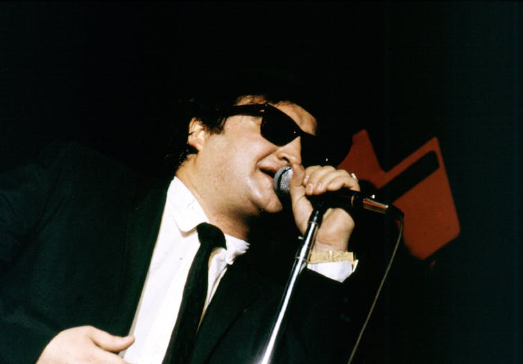 John Belushi in his Blues Brothers guise, wearing a black hat, black coat and tie and sunglasses and singing into a hand-held microphone.