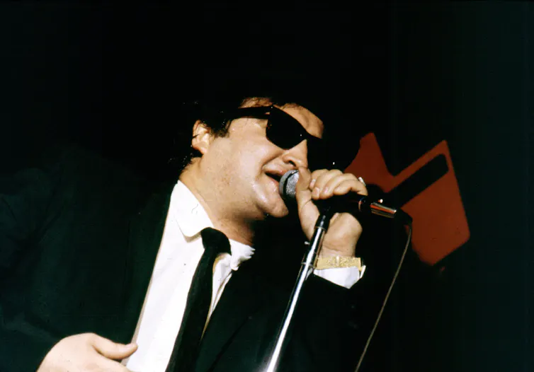 John Belushi in his Blues Brothers guise, wearing a black hat, black coat and tie and sunglasses and singing into a hand-held microphone.
