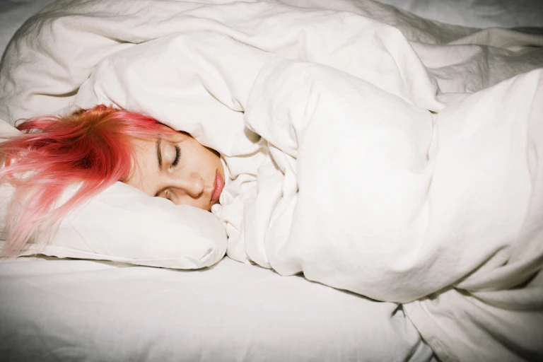 Young woman with bright red-orange dyed hair wrapped sleeping in a white duvet.