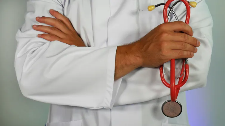 Photo of a person's torso in a white coat, which arms folded, holding a red stethoscope