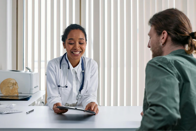 A woman in a white coat and stethoscope sitting across from a man with a ponytail, seen from behind.