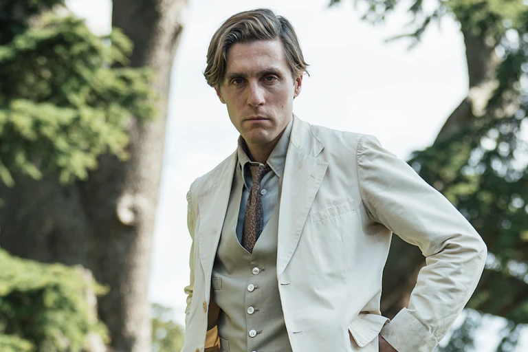 A sad and dishevelled-looking man staring at the camera well in a cream linen suit, waistcoat and tie in a 1940s style.