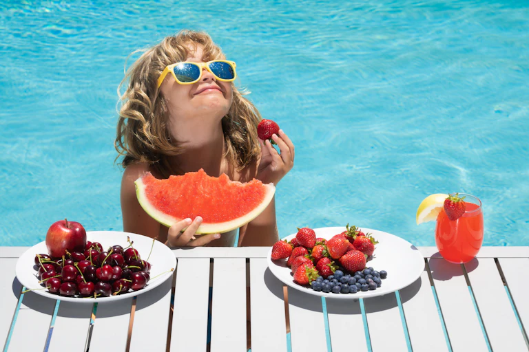 A smiling child wearing a yellow pair of sunglasses stands in a pool, eating a large watermelon slice and some strawberries.