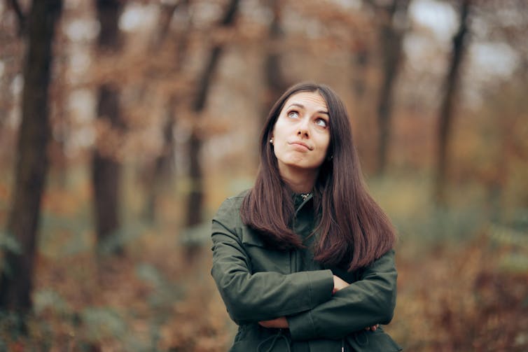 A woman wearing a green rain coat walks out in the autumn or winter forest, looking at the sky. She looks pensive or tired.