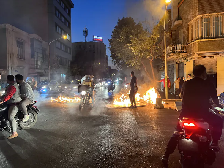 People of motorcycles drive in front of objects set alight on a street.