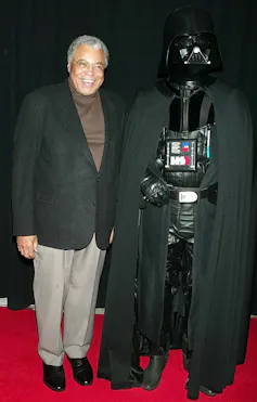 Older Black man wearing suit jacket poses on red carpet next to sinister-looking, costumed person wearing a black mask and black cape.