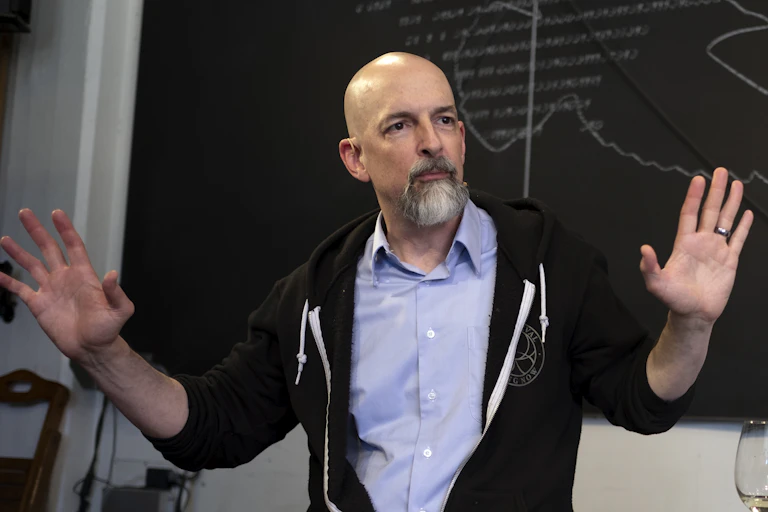 Photo of bald man with goatee sitting on a desk with both hands raised.