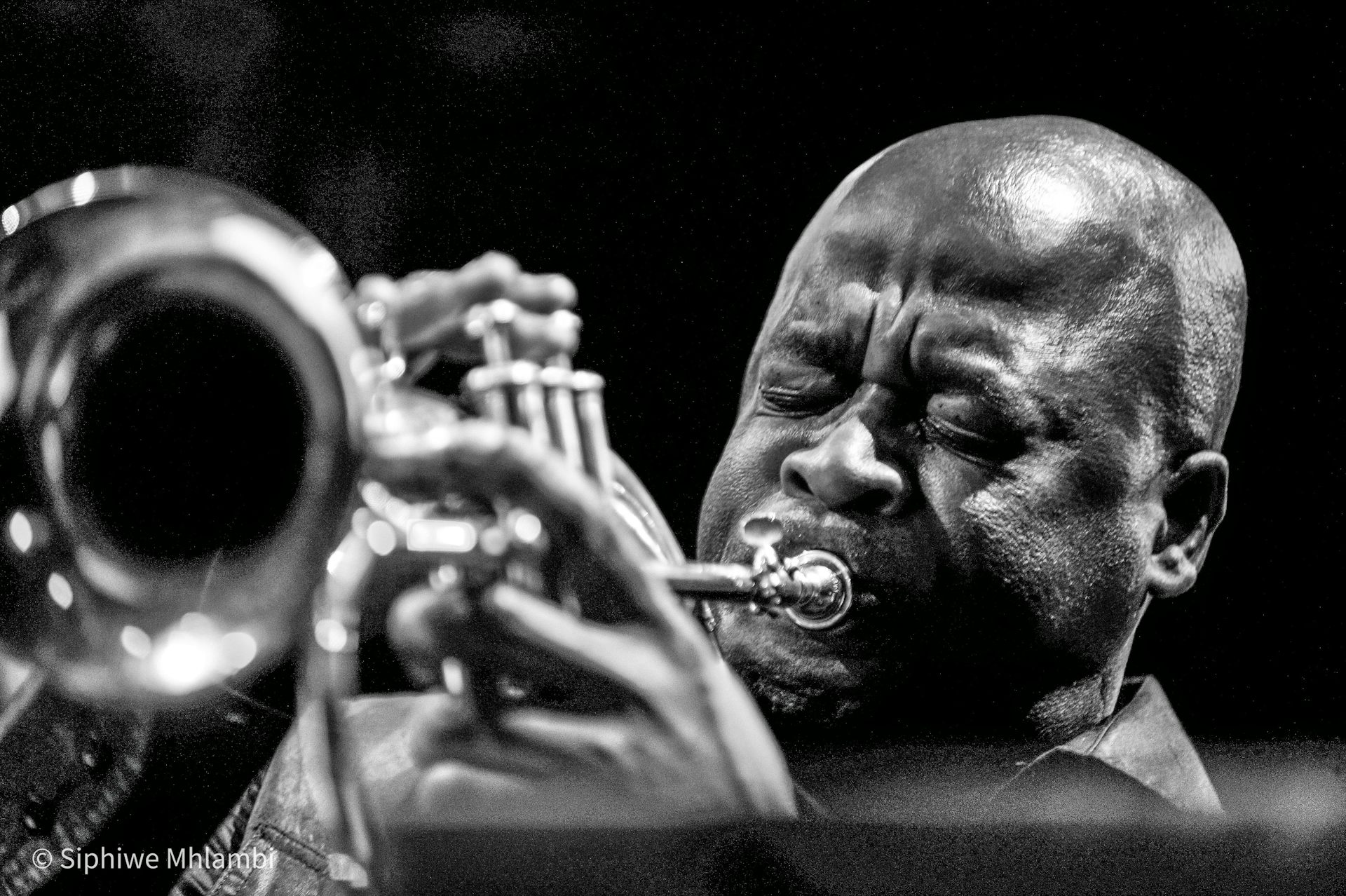 A black and white photo of a bald-headed African man blowing into a trumpet, his eyes closed.