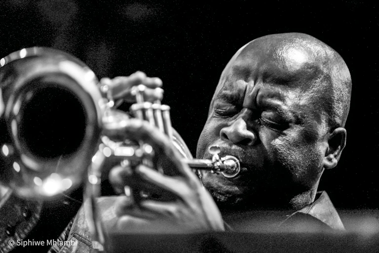A black and white photo of a bald-headed African man blowing into a trumpet, his eyes closed.