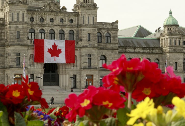 Una gran bandera canadiense en la entrada de un edificio de piedra con flores en primer plano.