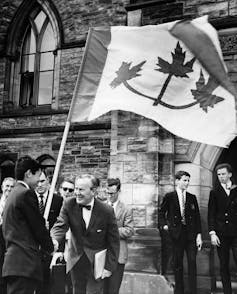 La foto en blanco y negro muestra a un adolescente que sostiene una bandera grande con tres hojas de arce y un hombre mayor en una cebolla de corbata sonriendo y estrechando su mano.