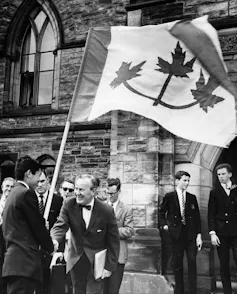 A black-and-white photo shows a teenager holding a large flag with three maple leaves and an older man in a bow tie smiling and shaking his hand.