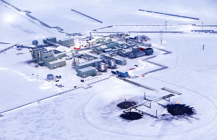 Industrial buildings in the snow have several roads and pipelines running to them and three wells with flares and blackened areas around them.